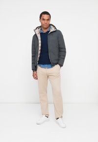 Giubbotto imbottito grigio con una leggera fodera beige, chiusura a zip e cappuccio. Indossato sopra un maglione blu navy e una camicia azzurro chiaro, abbinato a pantaloni beige e sneaker bianche.
