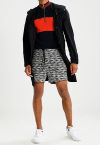 Homme portant une veste à capuche noire, un sweat à fermeture éclair noir et rouge, un short gris à motifs et des baskets blanches, se tenant devant un fond uni.