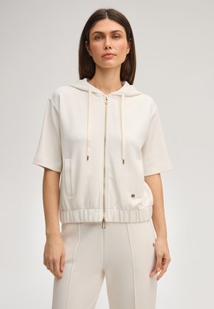 Femme portant un sweat à capuche crème à manches courtes avec fermeture éclair et un pantalon crème assorti, debout devant un fond clair uni.