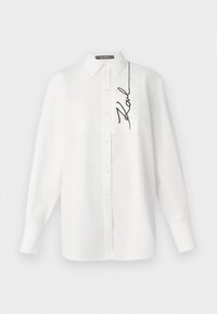 Camicia bianca a maniche lunghe in cotone, con tasca frontale e firma ricamata nera sul lato sinistro. Colletto classico e chiusura con bottoni.