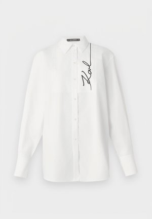 Bijela košulja dugih rukava od pamuka, s prednjim džepom i crnim izvezenim potpisom na lijevoj strani. Klasični ovratnik i kopčanje na gumbe.