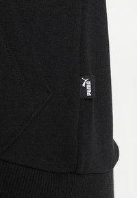 Tissu de sweat-shirt noir, avec des accents côtelés, une poche kangourou, et une petite étiquette logo Puma blanche sur la couture latérale.