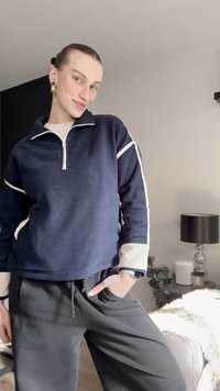 Marineblauwe fleece trui met witte rits en zijstrepen; gecombineerd met zwarte broek. Heeft lange mouwen en een ontspannen pasvorm.