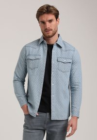 Lichtblauw denim overhemd met een subtiel patroon, drukknopen, twee borstzakken en lange mouwen, gedragen over een zwart shirt.