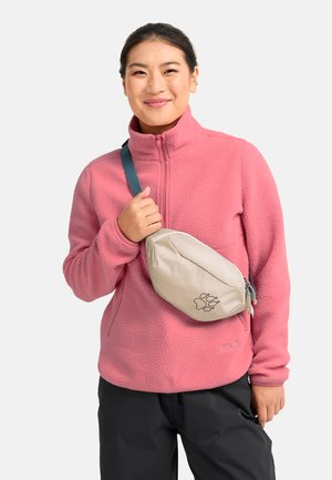 Glijdende persoon die een roze fleecejack en zwarte broek draagt, met een beige crossbodytas met een pootafdruklogo.