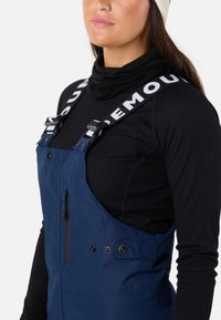 Blauwe waterdichte overall met verstelbare banden, voorzien van een wit logo, borstzak en drukknoopsluitingen. Draag dit over een zwarte, strak aansluitende turtleneck.
