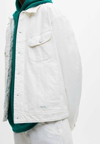 Vit denimjacka med avslappnad passform, som har två framfickor, metallic knappar och ett lätt tyg. Buren över en grön hoodie.