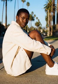 Persona ar īsiem matiem, vilkdama gaišu jaku un baltas sporta kurpes, sēž uz ietves, saulrietā fonā redzamas palmas un iela.