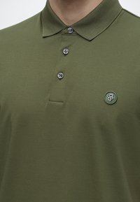 Homme portant un polo vert olive avec trois boutons et un petit logo rond sur la partie gauche de la poitrine.