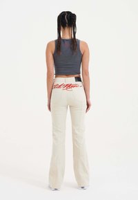 Pantalons évasés de couleur claire avec un texte brodé rouge à l'arrière et un patch. Associés à un haut sans manches gris, vus de l'arrière.