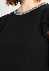 Black blouse featuring a rounded neckline adorned with a silver beaded trim and soft ruffled sleeves made of textured fabric.