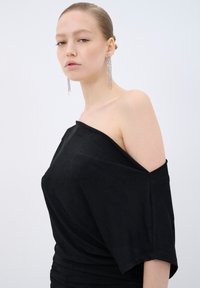 Sort off-shoulder top med et struktureret stof, der har løstsiddende ærmer og et samlet design. Model iført lange, glitrende øreringe.