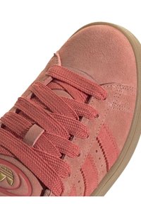 Rosa mockaläder sneaker med texturerade snören, rund tå och perforerade sidodetaljer. Sulan är ljusbrun med en slät yta.