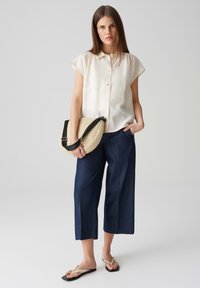 Helle beige Kurzarm-Hemd mit Knöpfen, marineblaue weit geschnittene Caprihosen, eine gewebte beigen Tasche mit einem schwarzen Tragegurt haltend, schwarze Flip-Flops tragend.