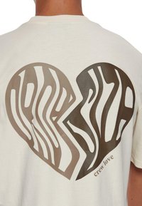 Cremfarbener Baumwoll-T-Shirt mit einem großen braunen Herzdesign mit wellenförmigen Streifen, dem Text "crew love" am unteren Rand, kurzen Ärmeln und normaler Passform.