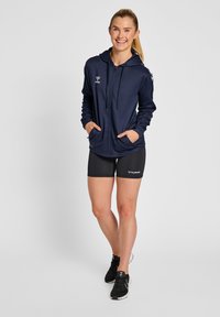 Giacca zip-up blu navy con cappuccio, maniche lunghe e tasche laterali. Indossata con pantaloni corti neri e scarpe da allenamento. Presenta dettagli con logo.