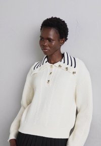 Pulover alb din tricot cu model tricotat, având o culoare cu dungi navy și butoane aurii pe plastron. Textura groasă a tricotului adaugă o dimensiune.