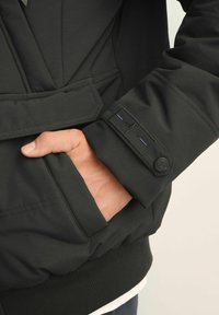 Czarna kurtka o gładkim, matowym wykończeniu. Posiada kieszeń na zamek błyskawiczny, ściągacze na rękawach oraz metkę na rękawie z informacjami o marce.