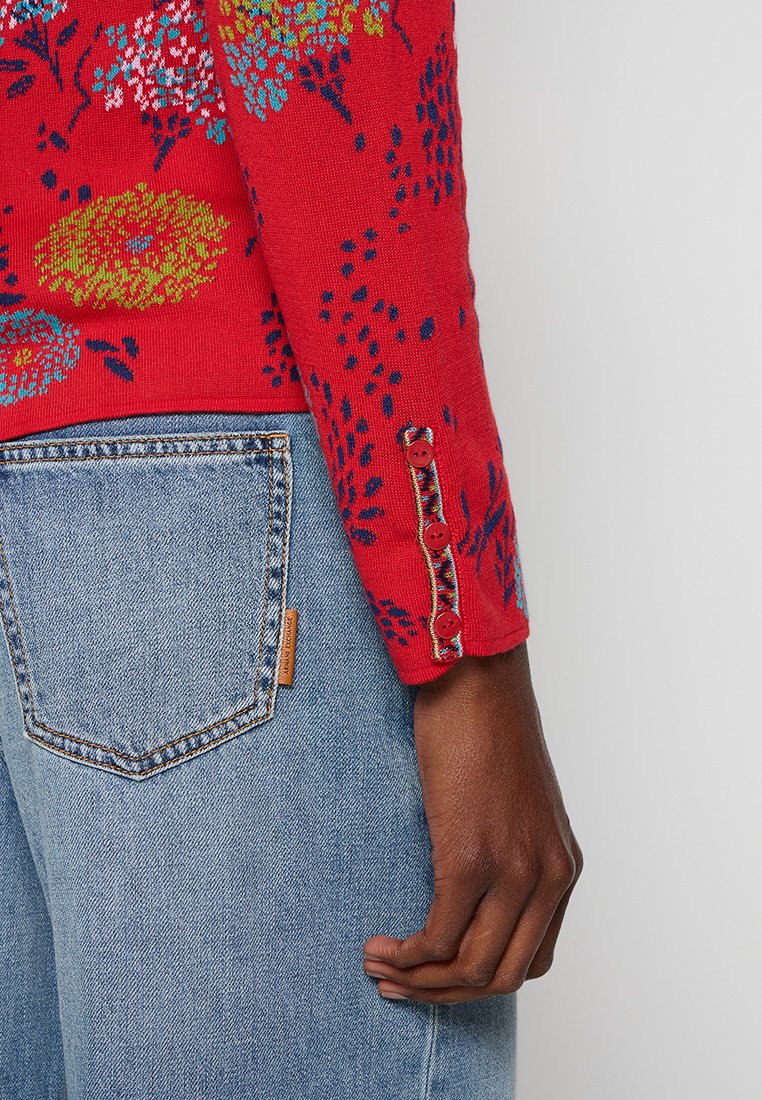 Czerwony sweter z dzianiny z kwiatowymi wzorami w niebieskim i żółtym, ozdobiony dekoracyjnymi guzikami przy mankietach, w zestawieniu z niebieskimi dżinsami.
