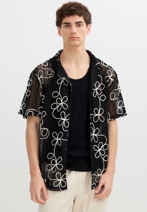 Joven con camisa negra de malla de manga corta con patrones florales blancos sobre una camiseta sin mangas negra y pantalones beige, de pie inmóvil.