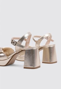 Sandalias de plataforma metálicas doradas con tacones de bloque; cuenta con correas en el tobillo ajustables y un diseño de dedos entrelazados. Textura suave y acabado brillante.