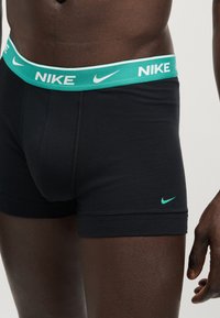 Μαύρα ανδρικά μποξέρ με πράσινη ζώνη που διαθέτει λευκό λογότυπο Nike και πράσινο σήμα Nike στο αριστερό μηρό, που φοριούνται από άτομο με σκουρόχρωμο δέρμα.