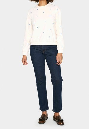 Person trägt weißes Sweatshirt mit kleinen blauen Herzen, dunkelblaue Jeans und braune Lederschlupfschuhe und steht vor einfachem Hintergrund.