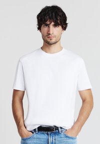 Uomo con capelli scuri che indossa una maglietta bianca a maniche corte e jeans blu chiaro, in piedi con le mani nelle tasche su uno sfondo neutro.
