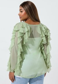 Blusa verde menta con tessuto trasparente, dettagli arricciati lungo le maniche e il collo, e una vestibilità ampia. Abbinata a jeans chiari.
