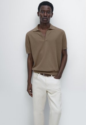 Modello maschile in piedi contro un muro chiaro, indossa una polo marrone a maniche corte, pantaloni bianchi e cintura marrone, con una mano in tasca.