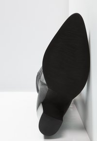 Zapato de tacón alto negro con punta afilada, material de ante y cuero, suela texturizada y diseño elegante sobre un fondo blanco.