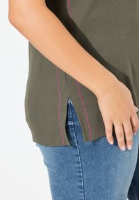 Olivgrünes T-Shirt mit gerippter Textur, ausgestattet mit seitlichen Schlitzen und kontrastierenden pinken Nähten. Kombiniert mit blauen Denim-Jeans.