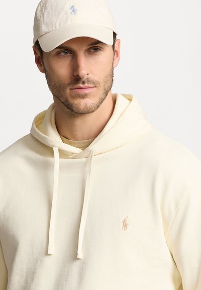 Krämfärgad hoodie med dragsnören och en liten ljusrosa polo-logotyp på bröstet. Bärs med en matchande ljusfärgad keps. Slät textur.