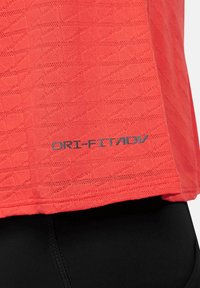 Punainen performance-paita, jossa on tekstuuri kuvio, harmaa DRI-FIT ADV -etiketti lähellä helmaa, yhdistetty mustiin leggingsseihin.