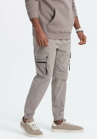 Man, der beigere Cargo-Hosen mit Reißverschluss- und Knopf-Taschen, beigen Sneakers und einem beigen Hoodie trägt, vor einem weißen Hintergrund steht.