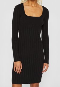 Robe noire côtelée à manches longues avec un décolleté carré. Elle présente une coupe ajustée et un tissu texturé avec un ourlet légèrement brut.