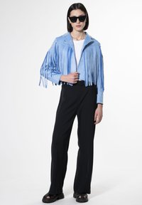 Giacca di suede azzurro chiaro con frange lunghe, abbinata a pantaloni neri a gamba larga e sandali neri spessi. Il modello indossa occhiali da sole scuri.
