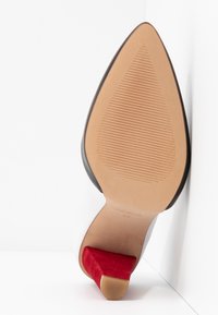 Semelle d’une chaussure à bout pointu avec une semelle inférieure beige texturée et un talon haut recouvert de suède rouge, incliné contre un coin blanc.