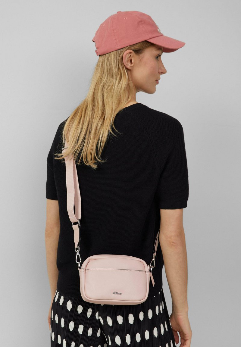 Rosa Umhängetasche aus strukturiertem Material, runde Form, verstellbarer Riemen, silberne Hardware, getragen über einem schwarzen Oberteil und gepunkteten Hosen.