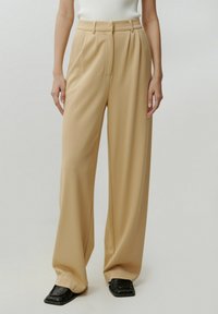 Beige wijde pantalon met een hoge taille, geplooide voorkant en een gladde textuur, gecombineerd met zwarte vierkante schoenen.