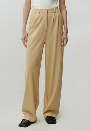 Beige Weite-Bein-Hosen mit hoher Taille, gefältelter Vorderseite und glatter Textur, kombiniert mit schwarzen eckigen Schuhen.