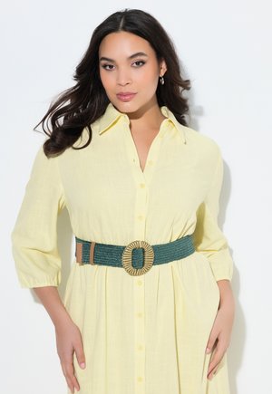 Donna con capelli lunghi e scuri che indossa un vestito giallo con bottoni e una larga cintura verde con fibbia rotonda intrecciata, in piedi davanti a uno sfondo bianco.