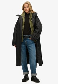 Lange zwarte gewatteerde jas over een groene shirt, gedragen met een zwarte trui en blauwe opgerolde jeans. Zwarte veterschoenen maken de look compleet.