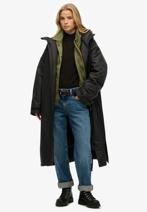 Lange zwarte gewatteerde jas over een groene shirt, gedragen met een zwarte trui en blauwe opgerolde jeans. Zwarte veterschoenen maken de look compleet.
