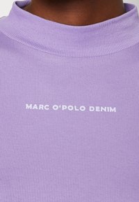 Detailní záběr na levandulově fialový žebrovaný top s bílým nápisem "MARC O'POLO DENIM" na hrudi a límečkem na krku.