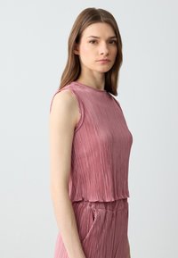 Top senza maniche di colore rosa con texture plissettata e scollatura rotonda. Il tessuto è leggero, creando una vestibilità comoda senza cuciture definite.
