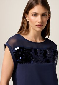 Camiseta de color azul marino con mangas cortas; presenta una capa superior transparente y una sección con lentejuelas en el pecho con lentejuelas oscuras y brillantes.