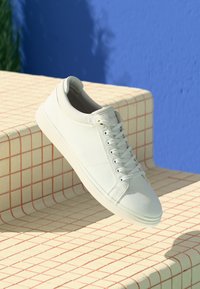 Baskets blanches en cuir texturé avec un bout lisse, des lacets ronds et une semelle en caoutchouc légère, posées sur une surface à motif en grille.