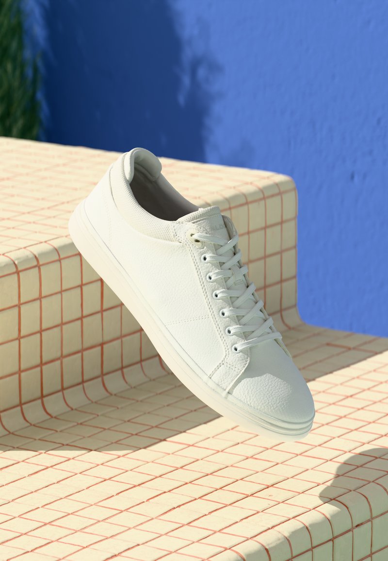 Baskets blanches en cuir texturé avec un bout lisse, des lacets ronds et une semelle en caoutchouc légère, posées sur une surface à motif en grille.