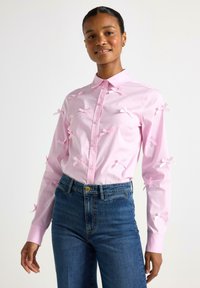 Camicia rosa con bottoni, adornata con piccoli fiocchi di raso sul davanti, abbinata a jeans blu a vita alta. Tessuto morbido, colletto classico, maniche lunghe.